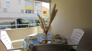 A dining area with a round table set for two, featuring a vase with dried pampas grass, a glass of water, and a plate with a knife and fork. The table is covered with a blue and yellow patterned tablecloth. There are two white chairs with green cushions. The room has a balcony with a view of a building and trees. - 40m² apartment ideal for 2 or 4.  50 m from the sea. quiet area. (Agde)