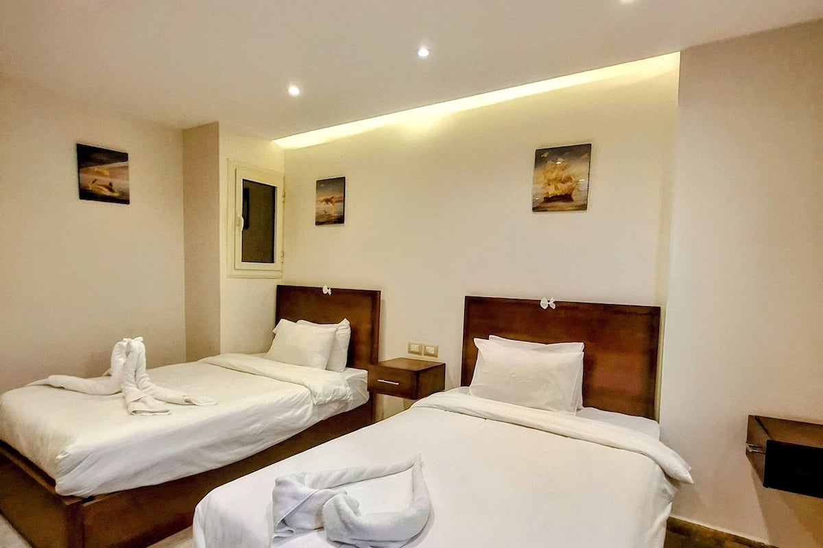 A hotel room with two beds, each with a wooden headboard and white bedding. There are wall-mounted paintings and a small bedside table with a lamp.