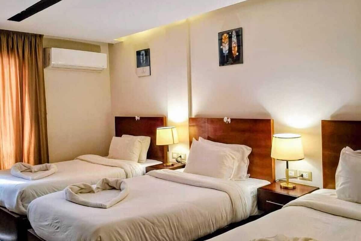 A hotel room with two beds, a wooden headboard, bedside lamps, an air conditioning unit, and a window with curtains.