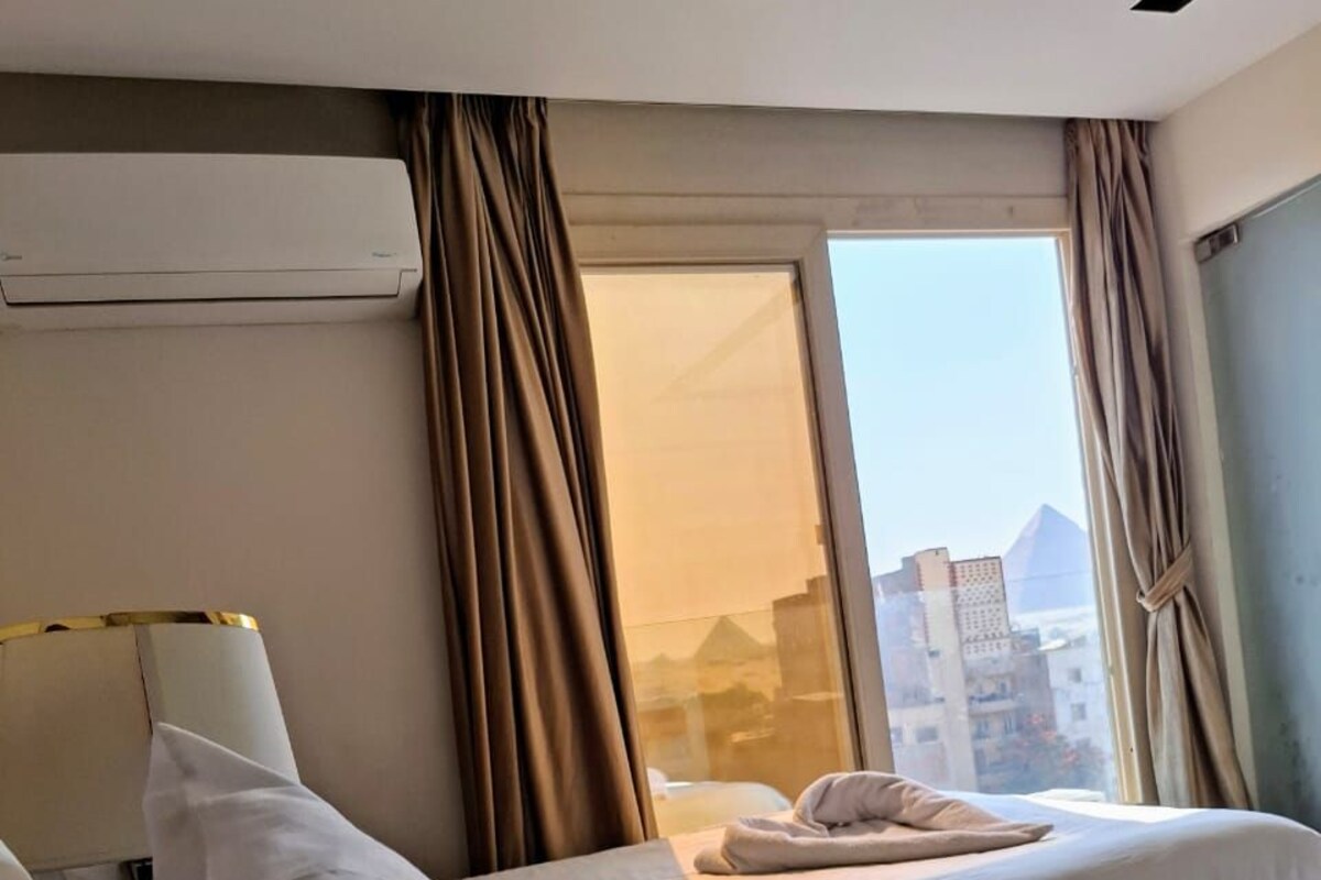 Une chambre d’hôtel avec deux lits, une fenêtre avec des rideaux, un climatiseur fixé au mur et une vue sur les bâtiments environnants.