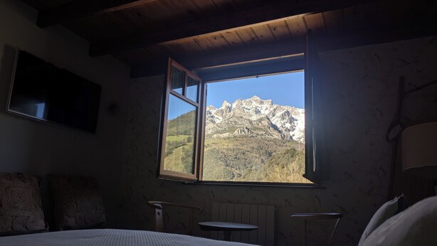A bedroom with a large window offering a mountain view, a bed, a small table, and a chair. - Luxury House in Picos de Europa Liebana garden with chill out area (La Frecha)