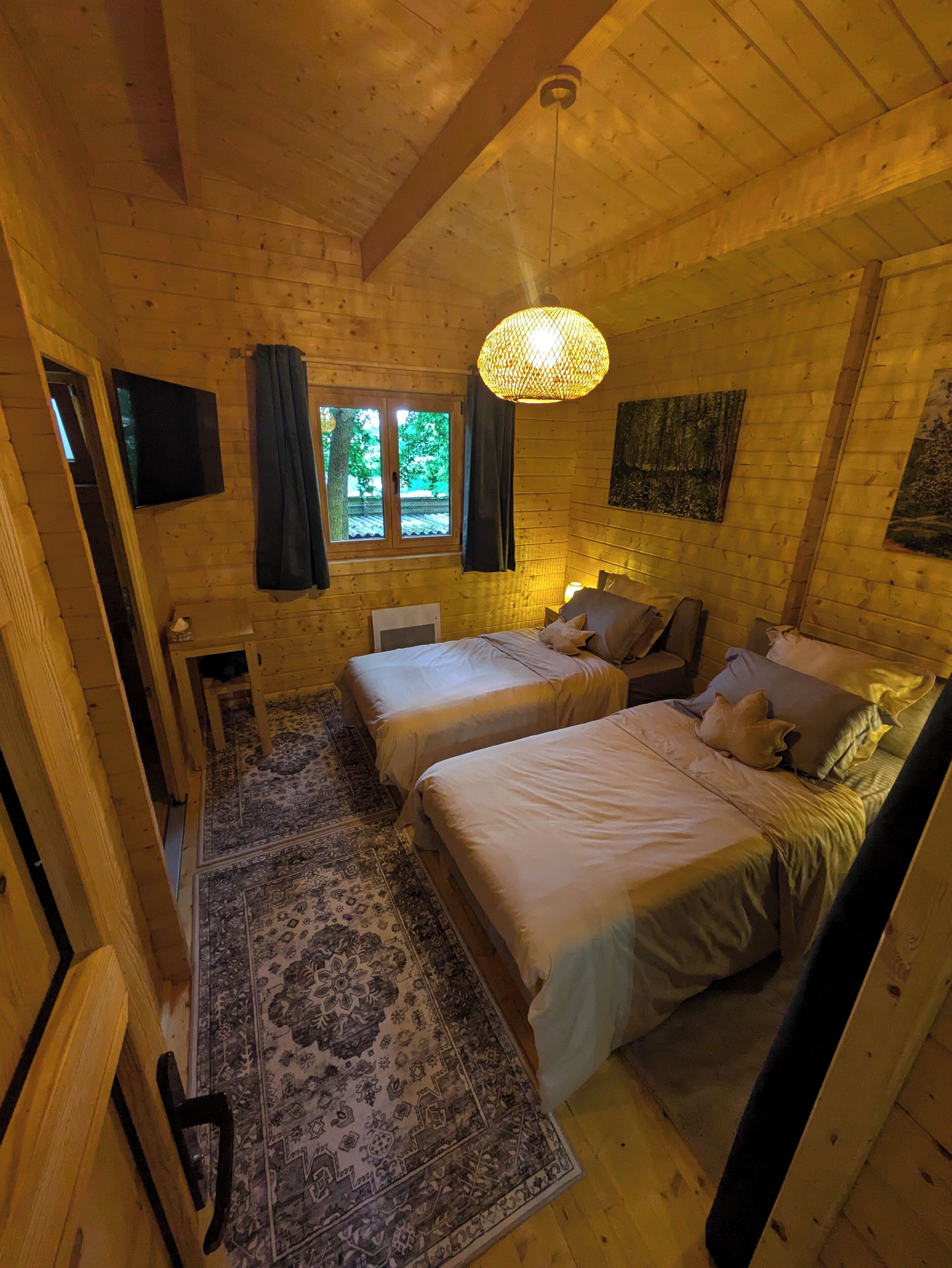 Une pièce chaleureuse, aménagée dans une cabane en bois, avec deux lits, un grand luminaire tressé, une fenêtre avec des rideaux et un téléviseur fixé au mur.