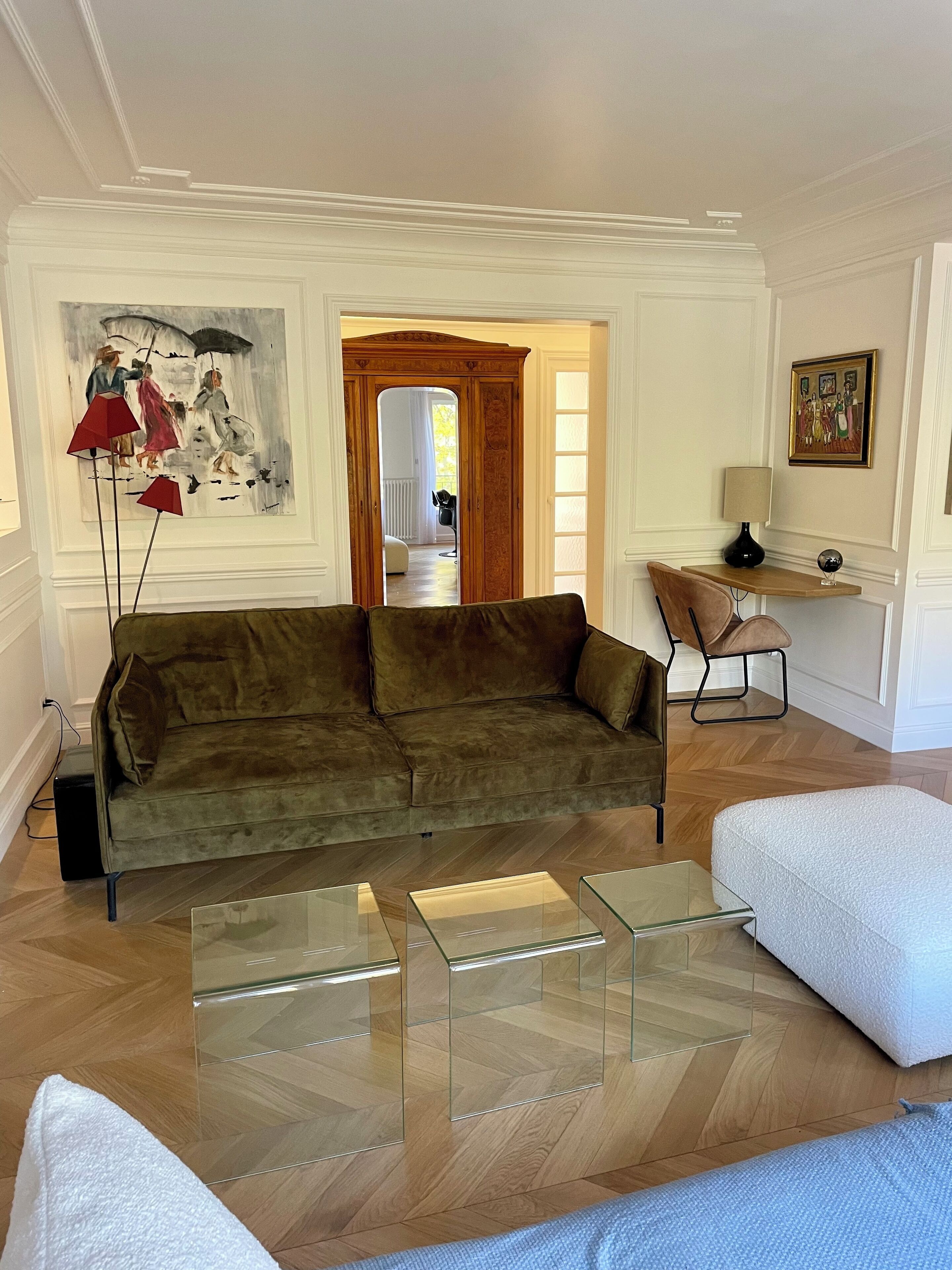 Ein Wohnzimmer mit einem grünen Sofa, einem Glastisch und einem weißen Sessel. An der Wand hängt ein Gemälde und auf einem Beistelltisch steht eine Lampe.
