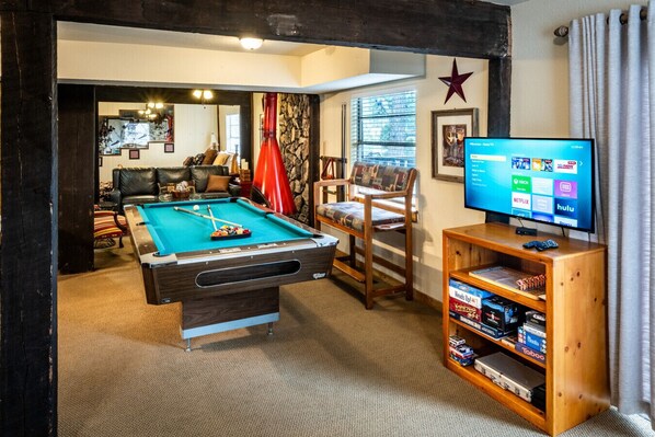 A cozy room with a pool table, a comfortable chair, a television, and a wooden shelf with video games. - Rocky Top - Condo in Midtown Ruidoso (Ruidoso)