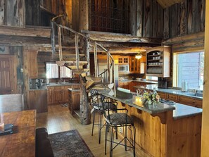 Een rustiek houten interieur met een wenteltrap, een keuken met houten kasten, een bar met krukken en een groot raam.