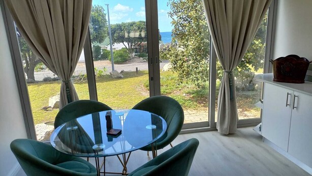 A modern room with a glass table and green chairs, large windows with curtains, and a view of a garden and trees. - Cosy apartment by the ocean, located in a family friendly town. (Hermanus)