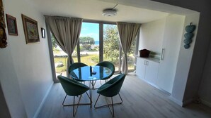 A dining area with a round glass table and four green chairs, a kitchen with white cabinets, and a view of the outdoors through large windows. - Cosy apartment by the ocean, located in a family friendly town. (Hermanus)