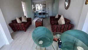 A modern living room with two sofas, a coffee table, and a glass-top dining table. There is a kitchen in the background with a window and a mirror on the wall. - Cosy apartment by the ocean, located in a family friendly town. (Hermanus)