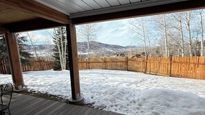 Une terrasse extérieure couverte, dotée d’un plancher en bois, d’une clôture en bois et offrant une vue sur un paysage enneigé, avec des arbres et des montagnes en arrière-plan.
