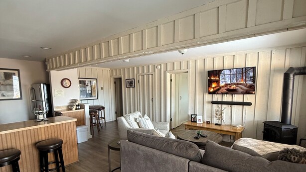 Un salon moderne avec un grand canapé d’angle gris, une table basse en bois et un téléviseur fixé au mur. On y trouve également un coin cuisine avec un bar et des tabourets, ainsi qu’une cheminée à l’arrière-plan.
