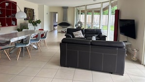 Un salón amplio con un sofá de cuero negro, una mesa de comedor con sillas, un televisor montado en la pared y un ventanal con vistas al exterior.