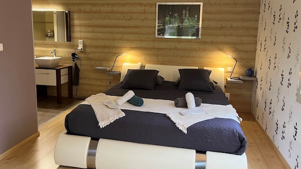 Un dormitorio moderno con pared de madera, cama con ropa oscura, dos lámparas de noche, un cuadro de paisaje urbano, una mesita de noche con espejo y papel tapiz estampado en la pared derecha.