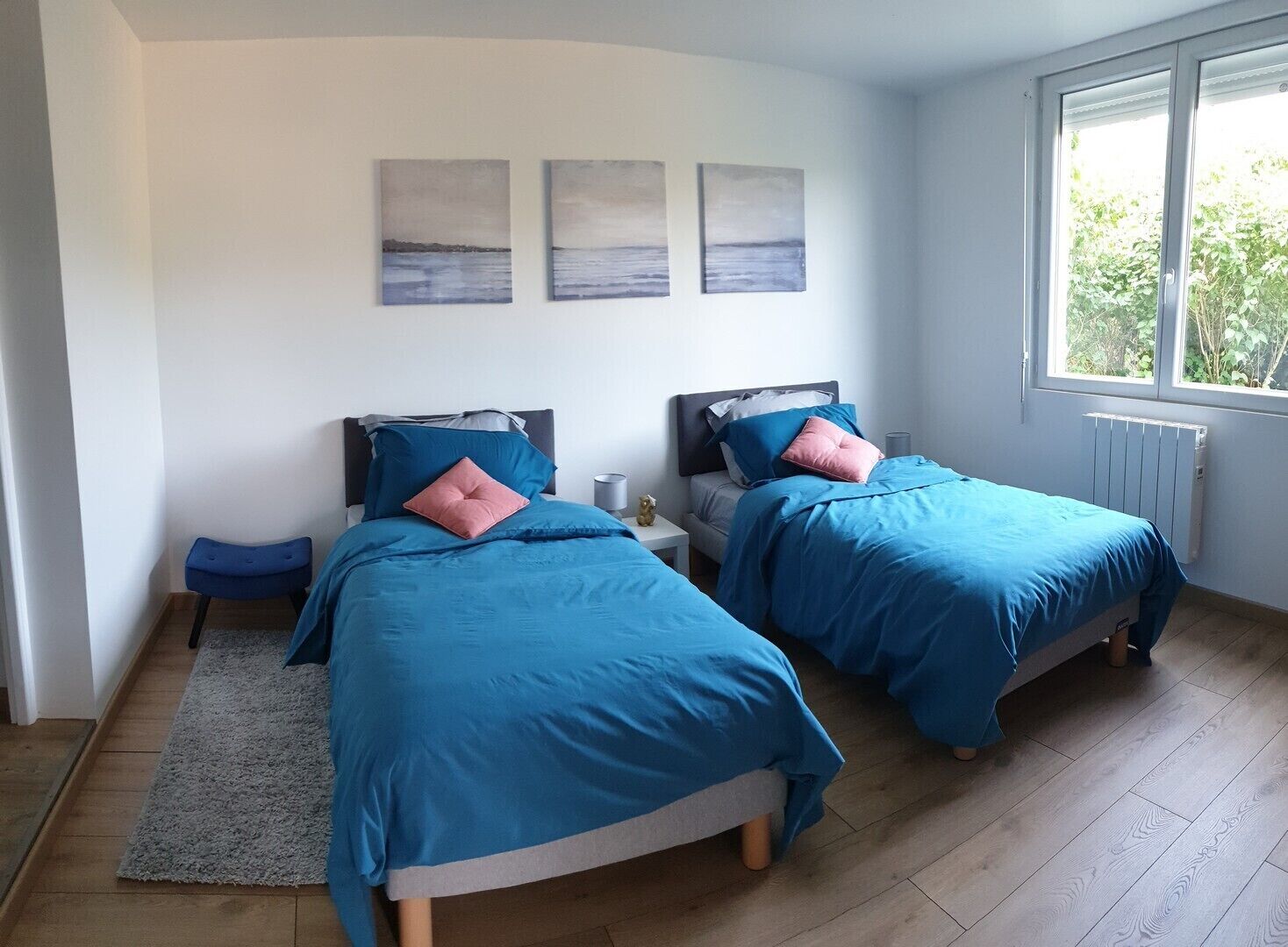 A bedroom with two beds, a grey rug, and a window with a view of greenery.