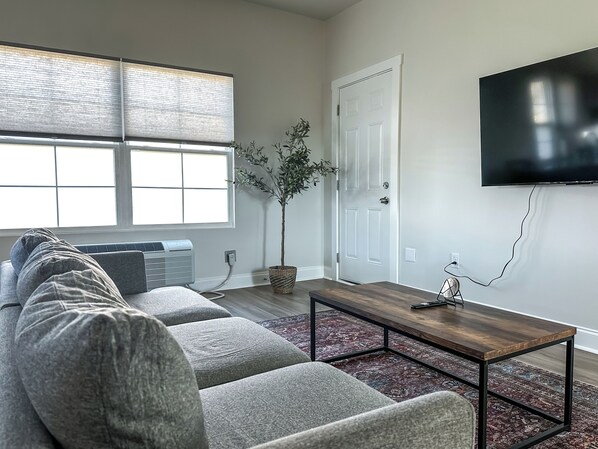 A modern living room with a grey sectional sofa, a wooden coffee table, and a large flat-screen TV mounted on the wall.