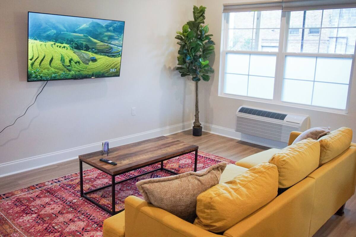 A living room with a flat-screen TV, a wooden coffee table, a yellow sofa, a red patterned rug, and a large window with a view of a building.