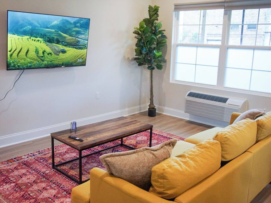 Una sala de estar con un televisor de pantalla plana, una mesa de centro de madera, un sofá amarillo, un tapete rojo estampado y un ventanal con vista a un edificio.