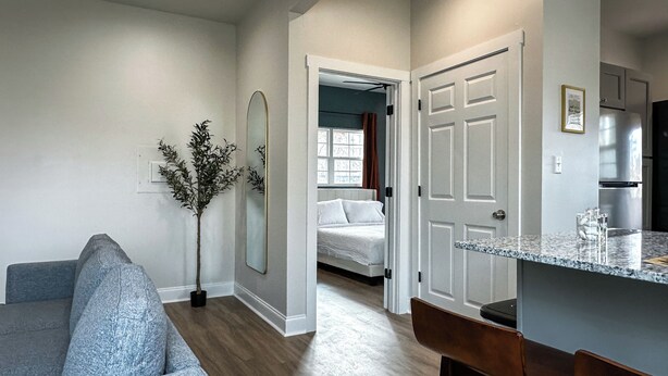 A modern living room with a grey sofa, a large mirror, and a potted plant. A bedroom with a bed and window is visible through an open door. - StayAE - Flamingo House (Baton Rouge)