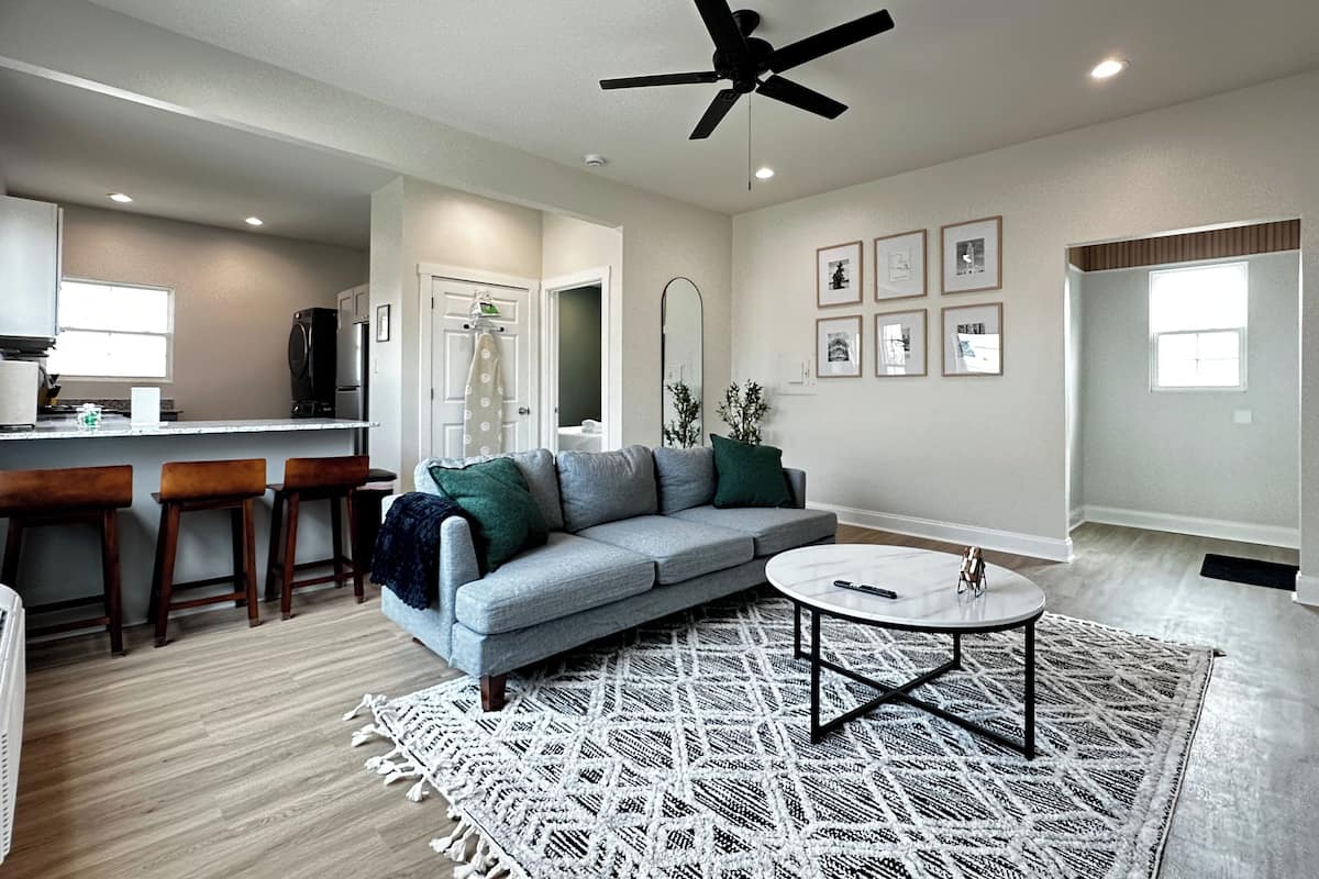 A modern living room with a grey sofa, a coffee table, and a ceiling fan. There is a kitchen in the background with a refrigerator and a window.