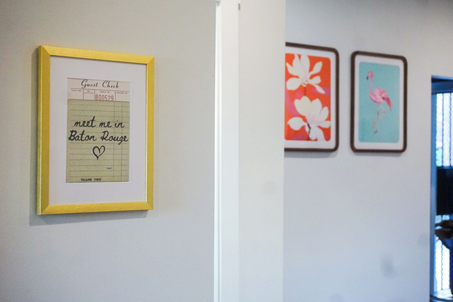 Un chèque de restaurant encadré, accompagné d’un mot manuscrit, une porte et deux œuvres d’art florales encadrées accrochées au mur.