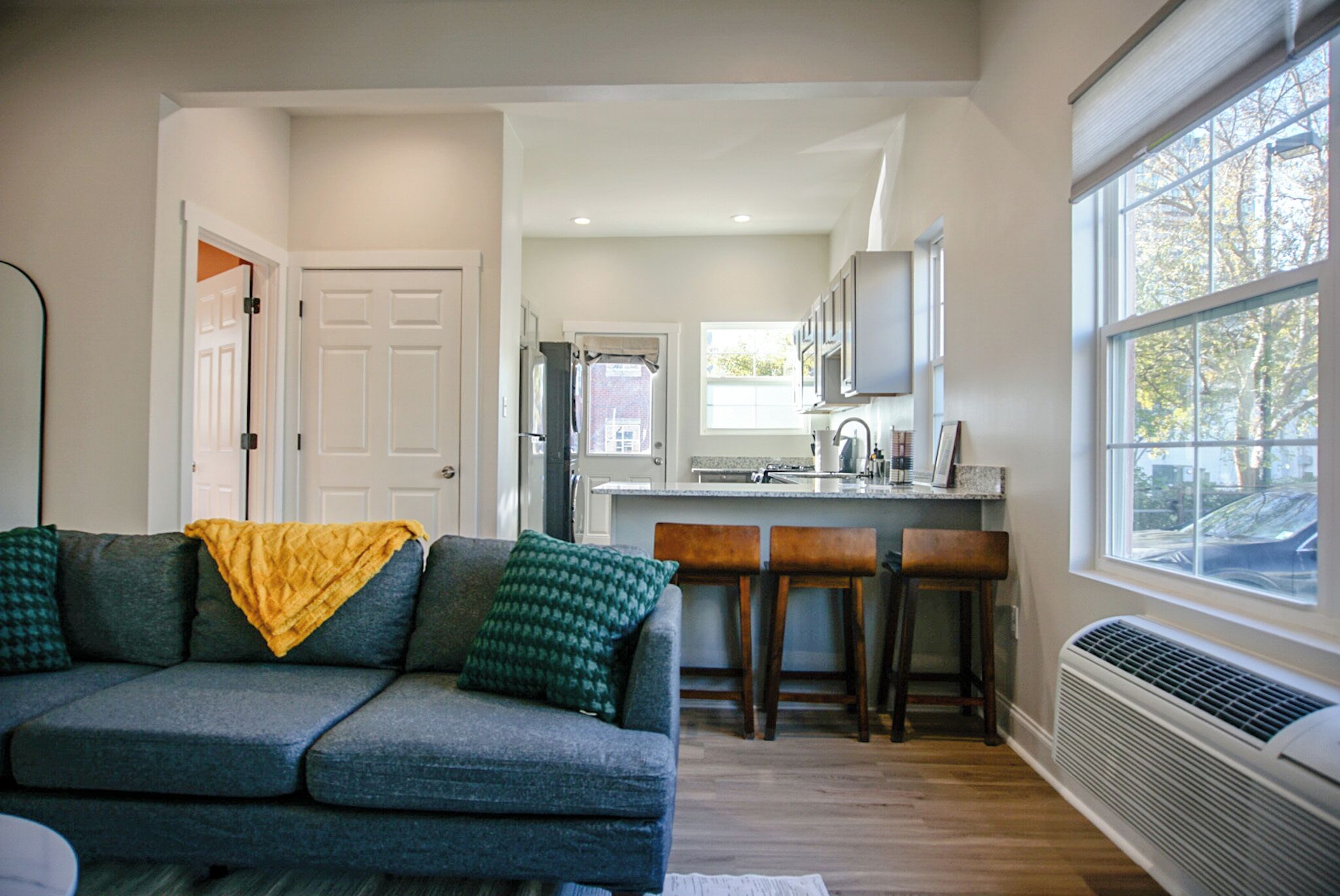 A modern living room with a gray sofa, a yellow throw blanket, and a green patterned pillow. There is a kitchen area with a white refrigerator, a microwave, and a window with a view of the outside.