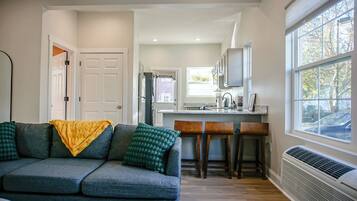 A modern living room with a gray sofa, a yellow throw blanket, and a green patterned pillow. There is a kitchen area with a white refrigerator, a microwave, and a window with a view of the outside.