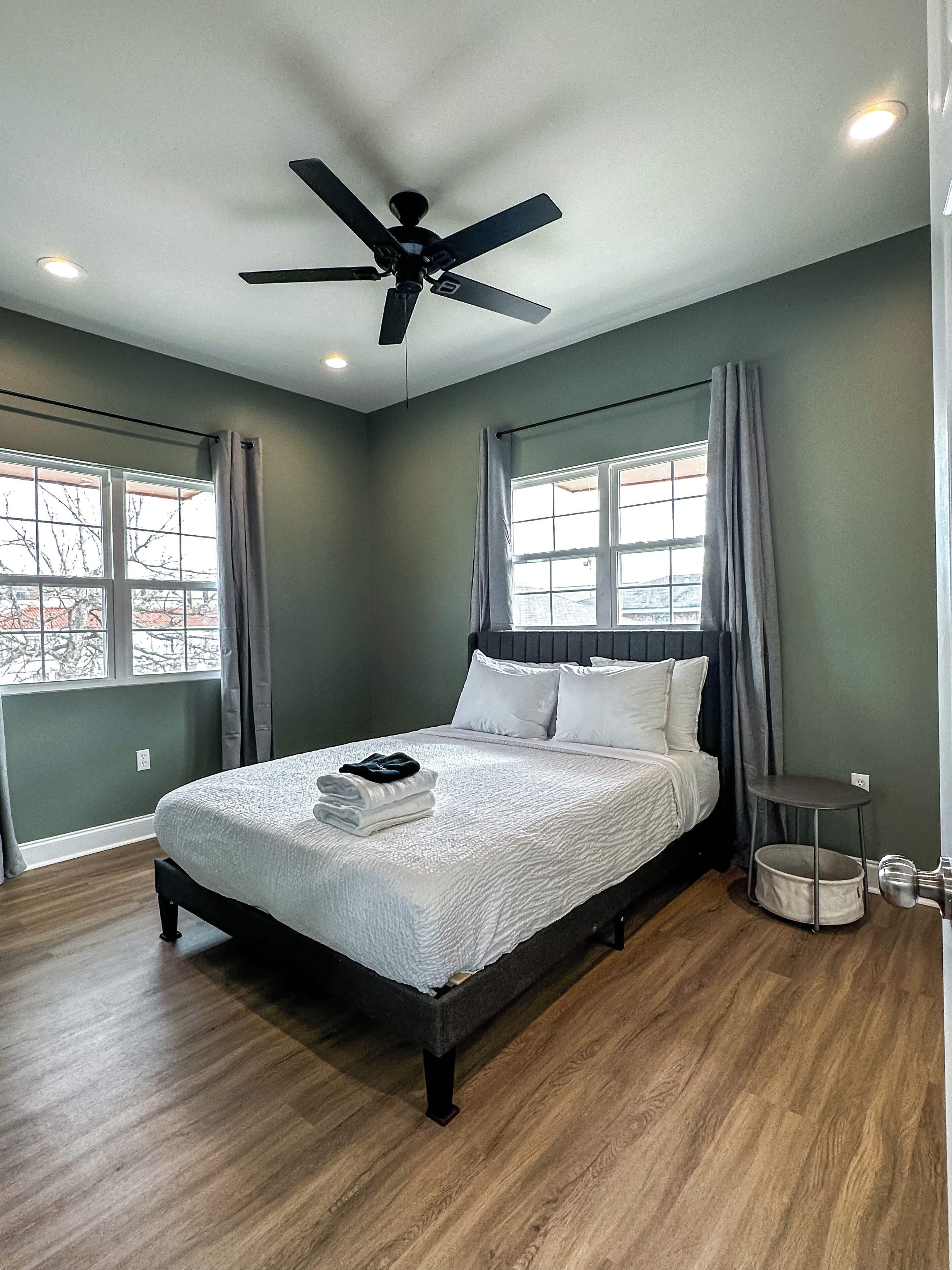 Un dormitorio con una cama, ventilador de techo, ventana con cortinas y una mesita de noche con lámpara.