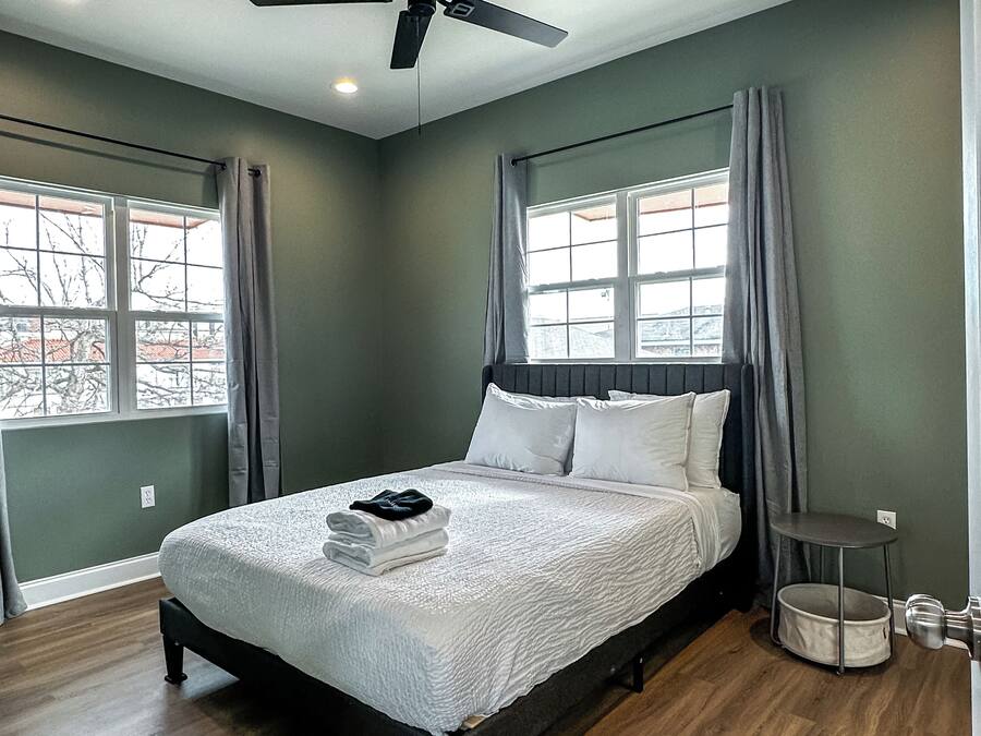 Un dormitorio con una cama, ventilador de techo, ventana con cortinas y una mesita de noche con lámpara.