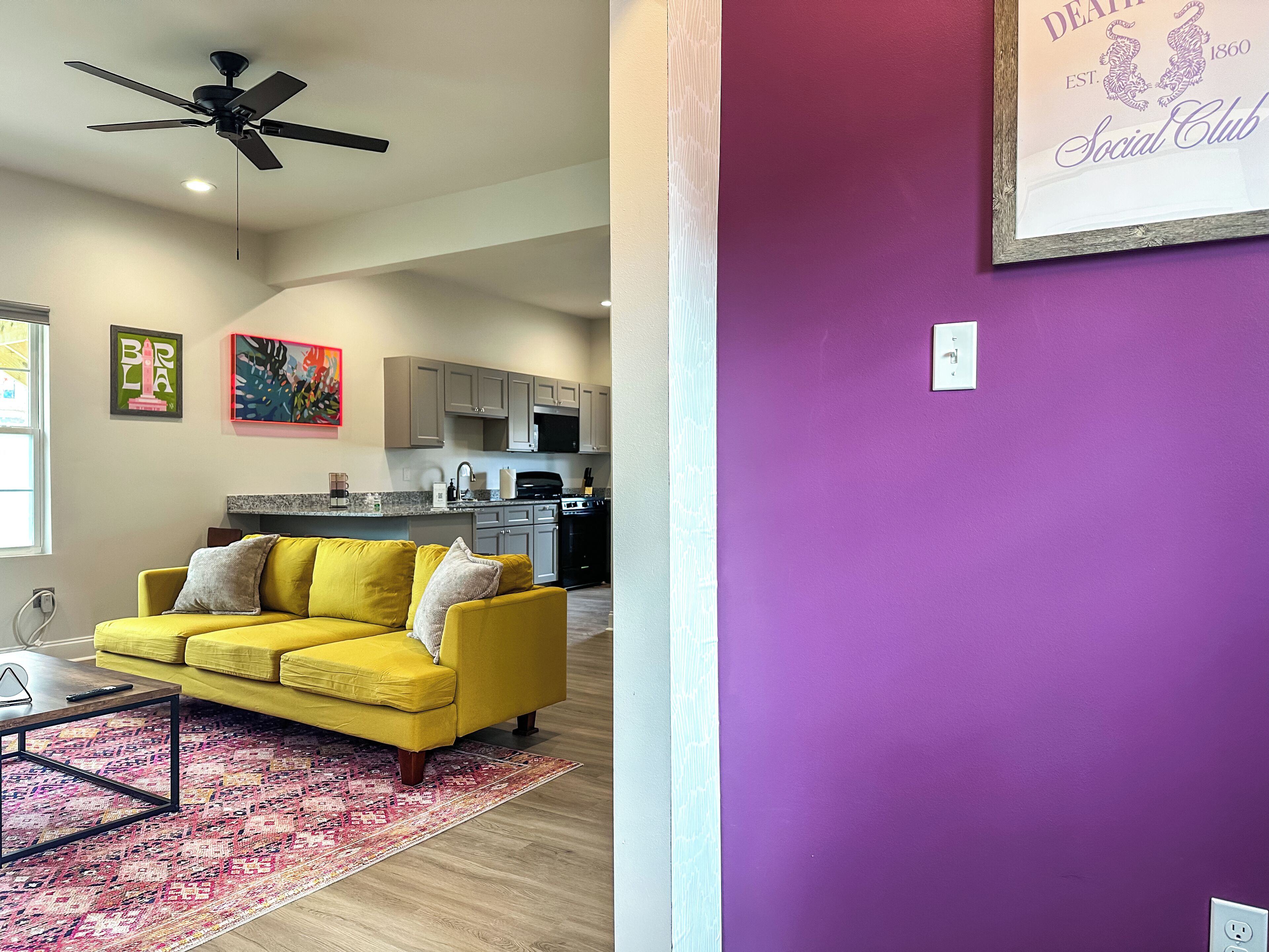 A living room with a yellow sofa, a ceiling fan, and a kitchen in the background.