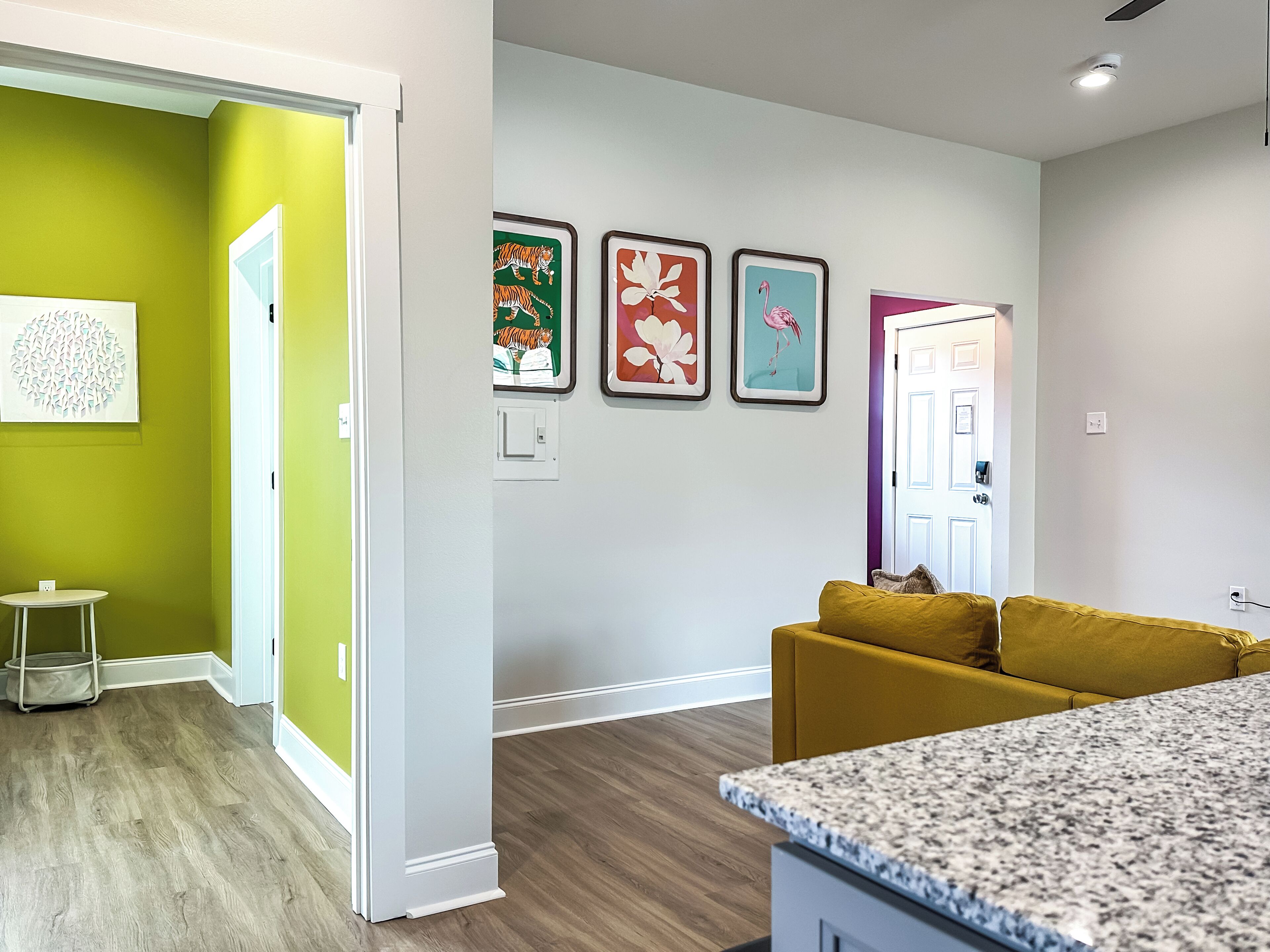 A modern living room with a yellow sofa, a kitchen area with granite countertops, and a hallway with green walls and artwork.