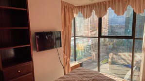 Ein Schlafzimmer mit einem Bett, einem Fernseher an der Wand und einem großen Fenster mit Vorhängen.