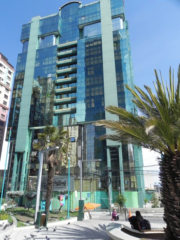 A modern building with a glass facade, surrounded by palm trees and a paved area with benches.