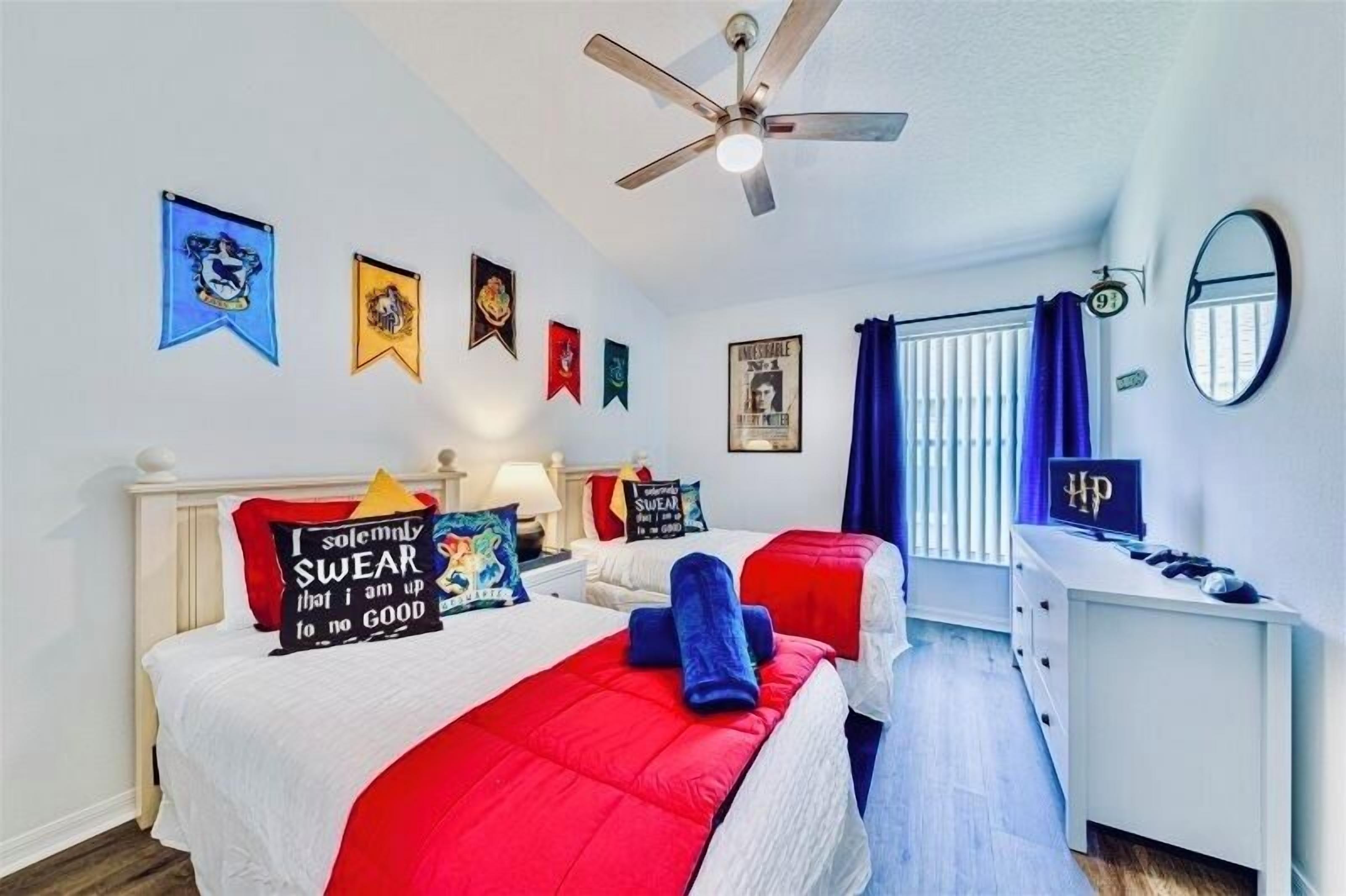 Quarto com duas camas, um ventilador de teto, uma escrivaninha com um laptop e diversas decorações nas paredes, incluindo bandeiras e cartazes.