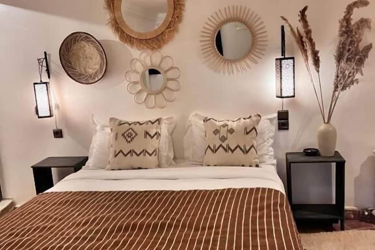 Un dormitorio con colcha a rayas, dos almohadas estampadas, un espejo redondo y apliques de pared.
