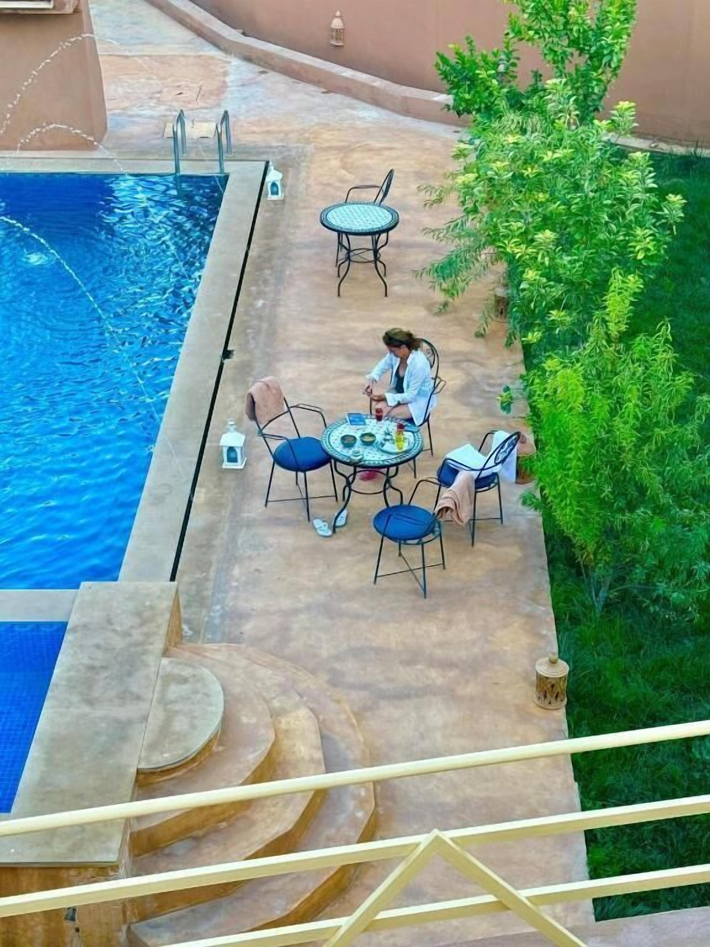 Zona junto a la piscina con una mesa, sillas, un pequeño árbol y vista despejada a la piscina.
