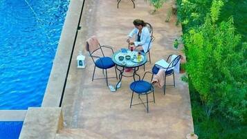 Zona junto a la piscina con una mesa, sillas, un pequeño árbol y vista despejada a la piscina.