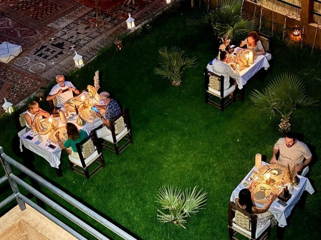 An outdoor dining area with tables and chairs arranged on a grassy lawn, surrounded by potted plants and decorative lanterns.
