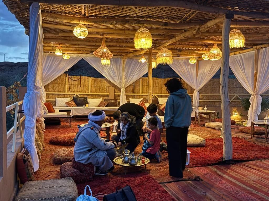 A tented area with multiple beds, hanging lights, and a group of people gathered around a table.