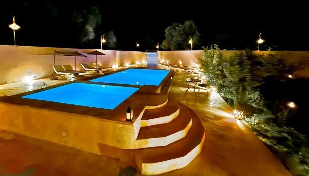 A nighttime swimming pool area with two pools, lounge chairs, and ambient lighting.