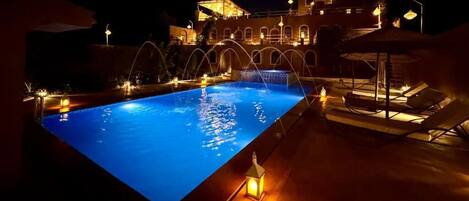 Une vue nocturne d’une piscine dont l’eau est illuminée, entourée de chaises longues et d’un bâtiment doté de fenêtres en arc de cercle.