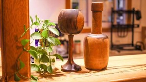 A wooden table with a plant and two wooden objects, possibly a cup and a bottle, in a room with a window and a chair in the background. - Evot Wave (Himmafushi)
