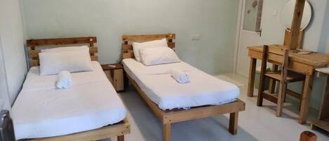 Dos camas de madera con sábanas y toallas blancas, un escritorio de madera y un espejo en una habitación con paredes de colores claros y suelo de baldosas.