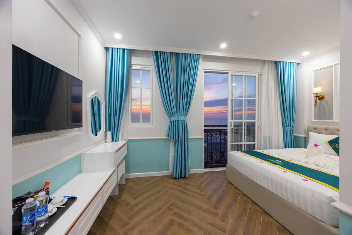 A hotel room with a bed, a vanity with a mirror, a window with curtains, and a balcony with a view of the sunset.