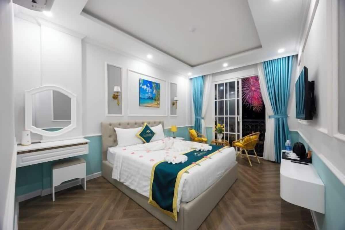 A modern hotel room with a bed, a vanity with a mirror, a desk, a chair, and a TV mounted on the wall.