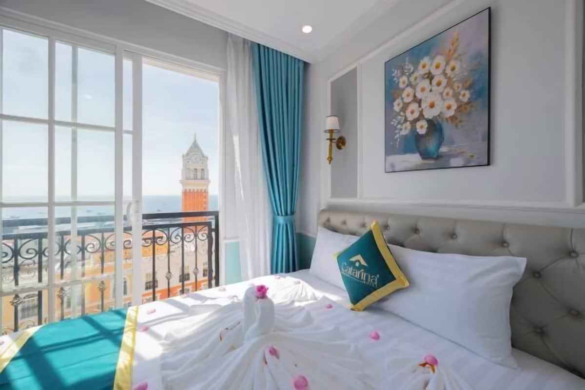 A hotel room with a bed, a painting, a view of a tower, and a balcony.