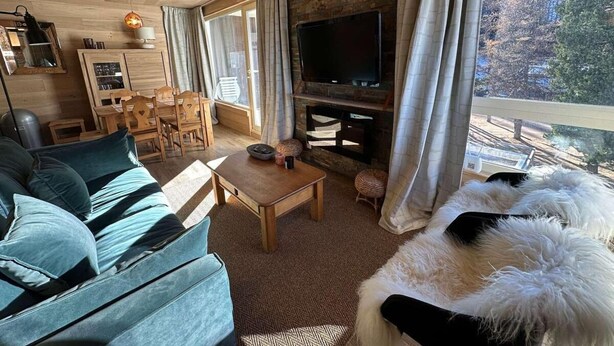 A cozy living room with a sofa, a coffee table, and a TV mounted on a stone wall. There is a dining area with chairs and a table, and a view of trees through the window. - 50m from ski slopes, 3rd floor, balcony, fireplace or stove, ski locker, 40m², Vars (Vars)