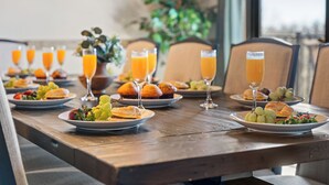 Une table à manger avec des assiettes remplies de nourriture, des verres de jus d’orange et une plante en arrière-plan.
