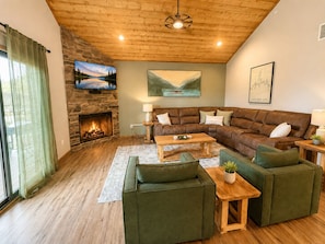 A cozy living room with a stone fireplace, a sectional sofa, a coffee table, and a television mounted on the wall. - 5BR w/ Home Theater, Hot Tub + Slip (Branson)