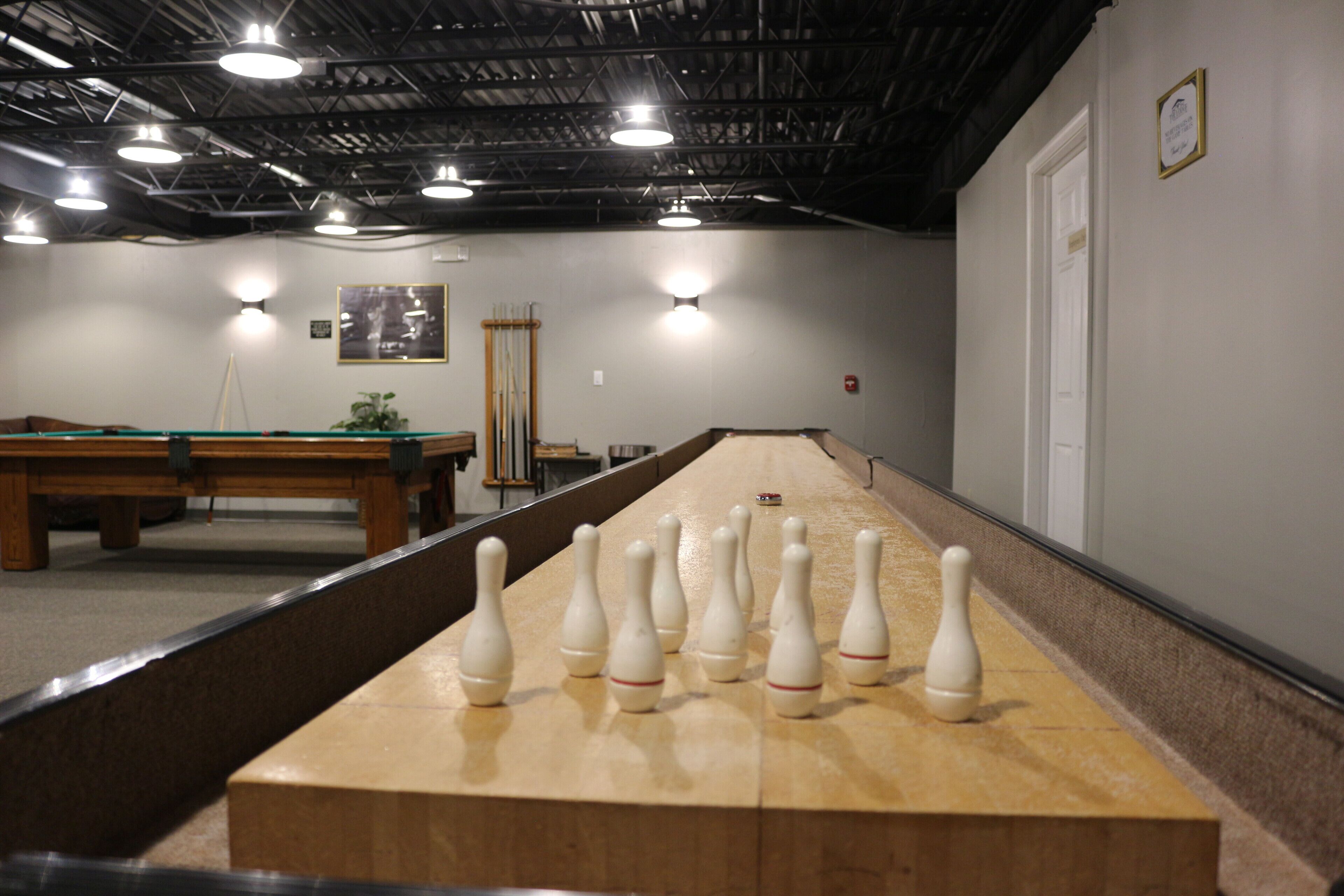 A long pool table with a dozen white bowling pins in the center, a billiards table in the background, and a high ceiling with industrial lighting.