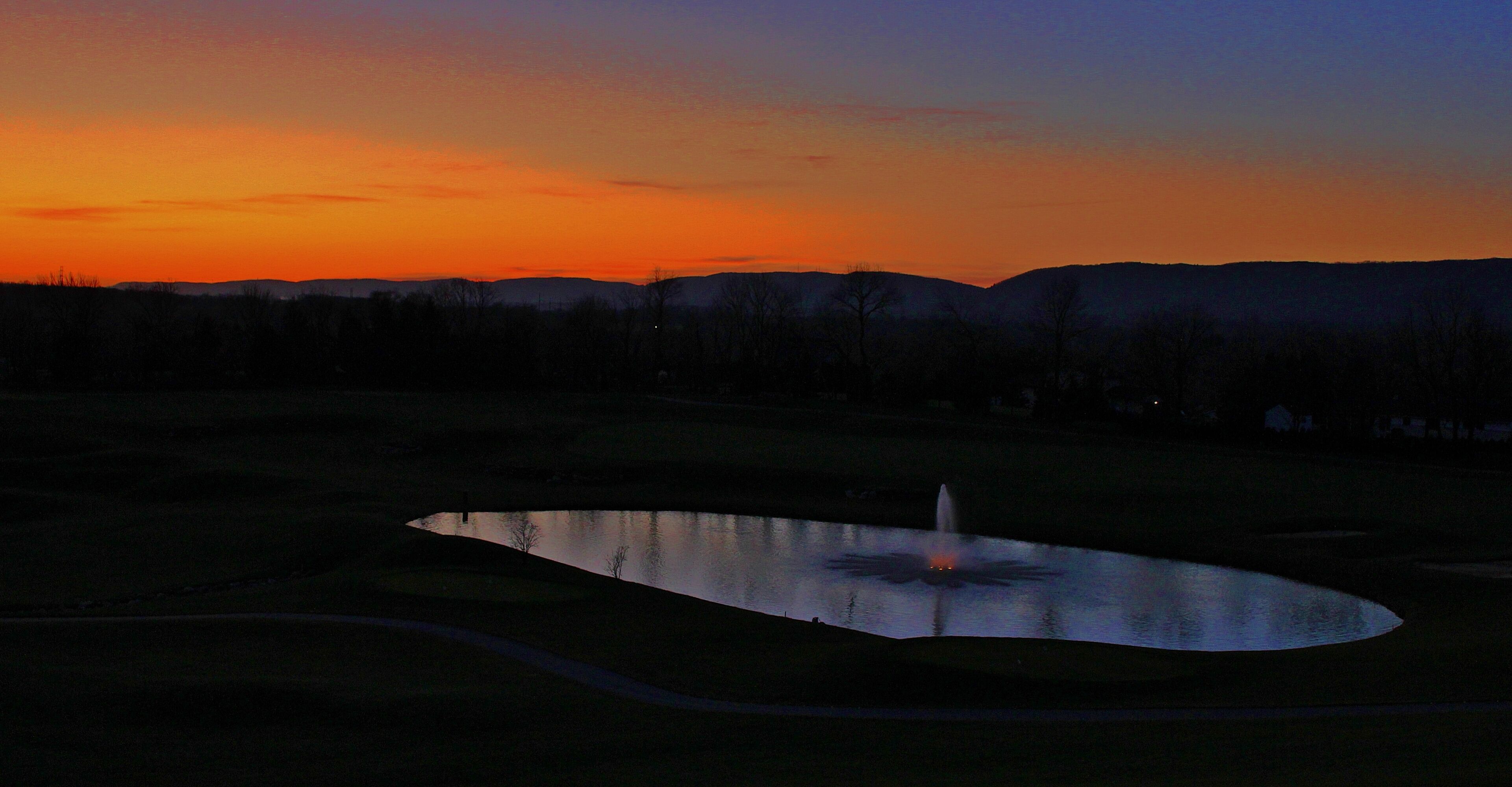 A serene landscape with a pond and a fountain, surrounded by hills and trees during sunset.