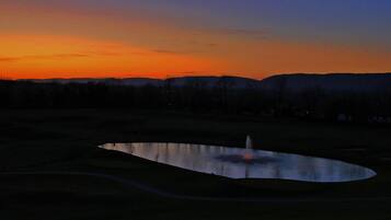 A serene landscape with a pond and a fountain, surrounded by hills and trees during sunset.